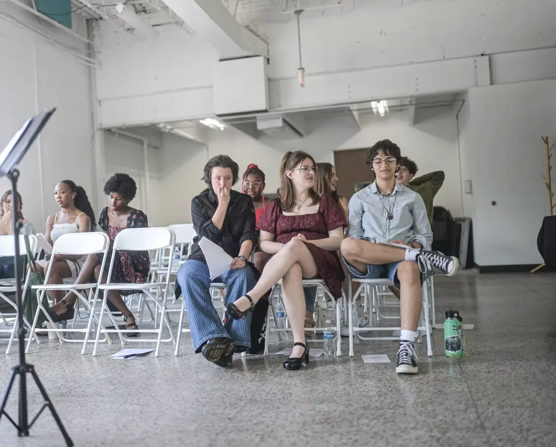 A row of teenagers seated on folding chairs in a bright studio space, listening.