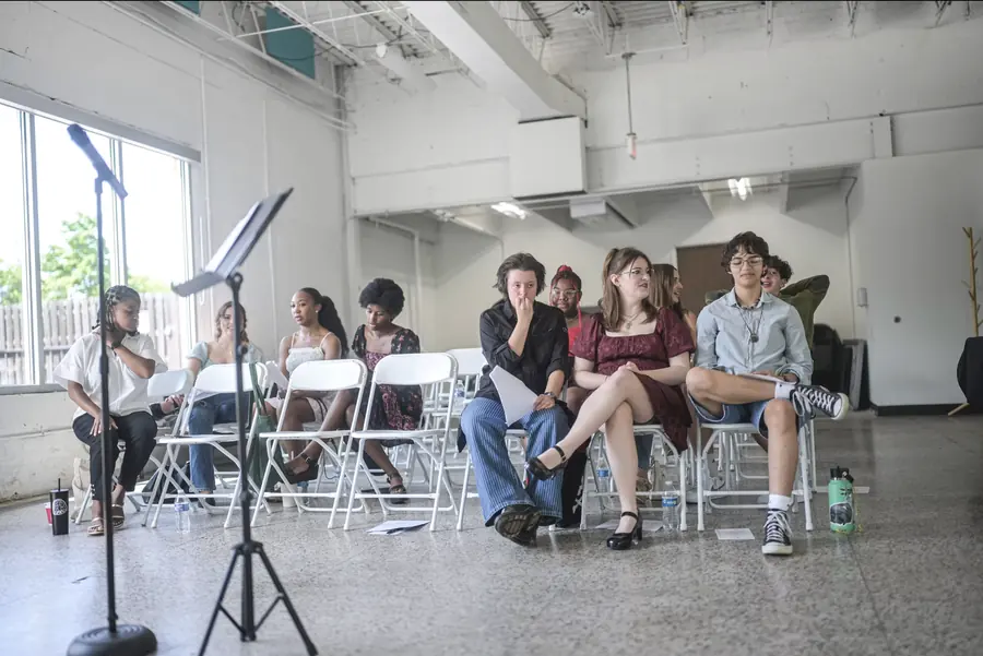 A row of teenagers seated on folding chairs in a bright studio, listening to a reading.