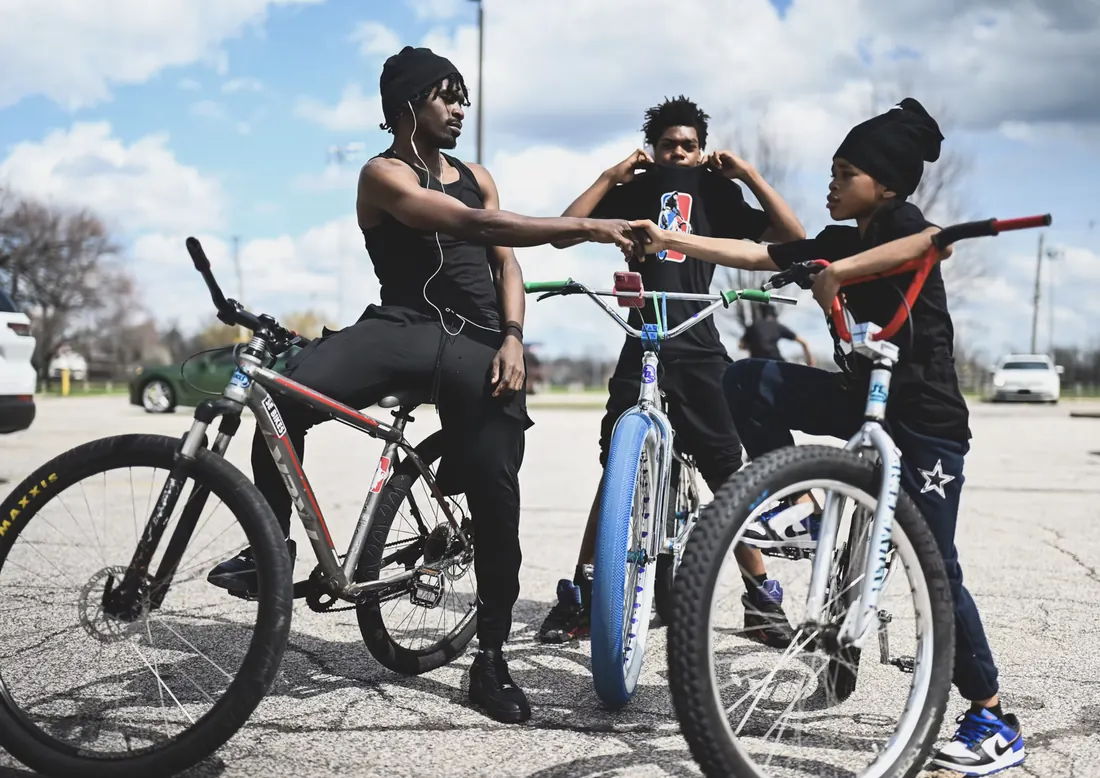 Three riders on customized bikes in a parking lot under a bright cloudy sky, two bumping fists.