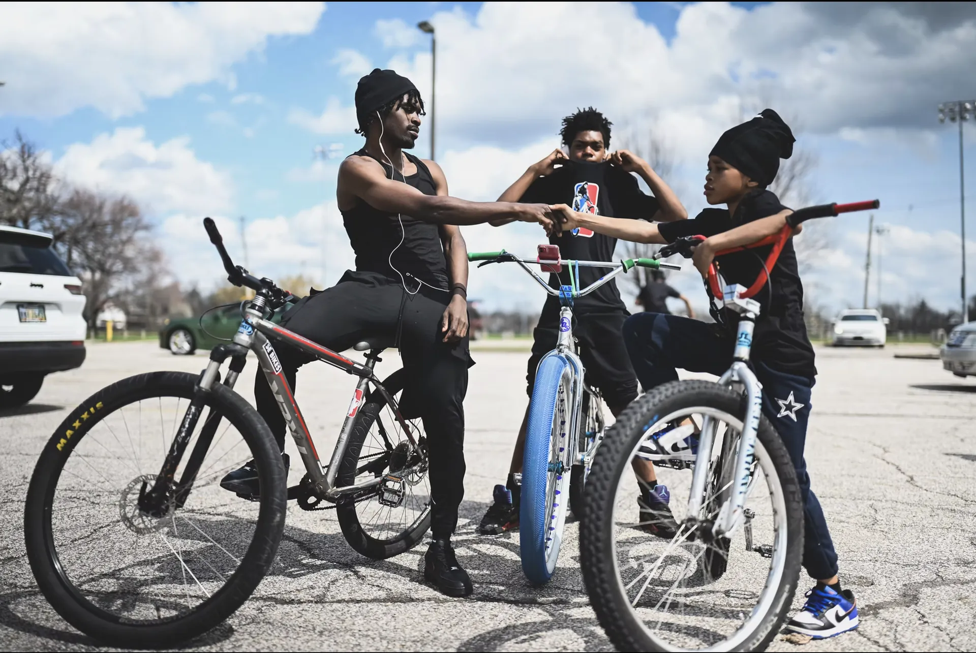 Three young riders on customized bikes in a parking lot under a bright sky, two of them bumping fists.