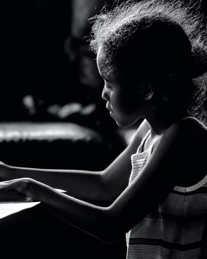A young girl drawing with a pencil at a table, lit from one side in a darkened room.