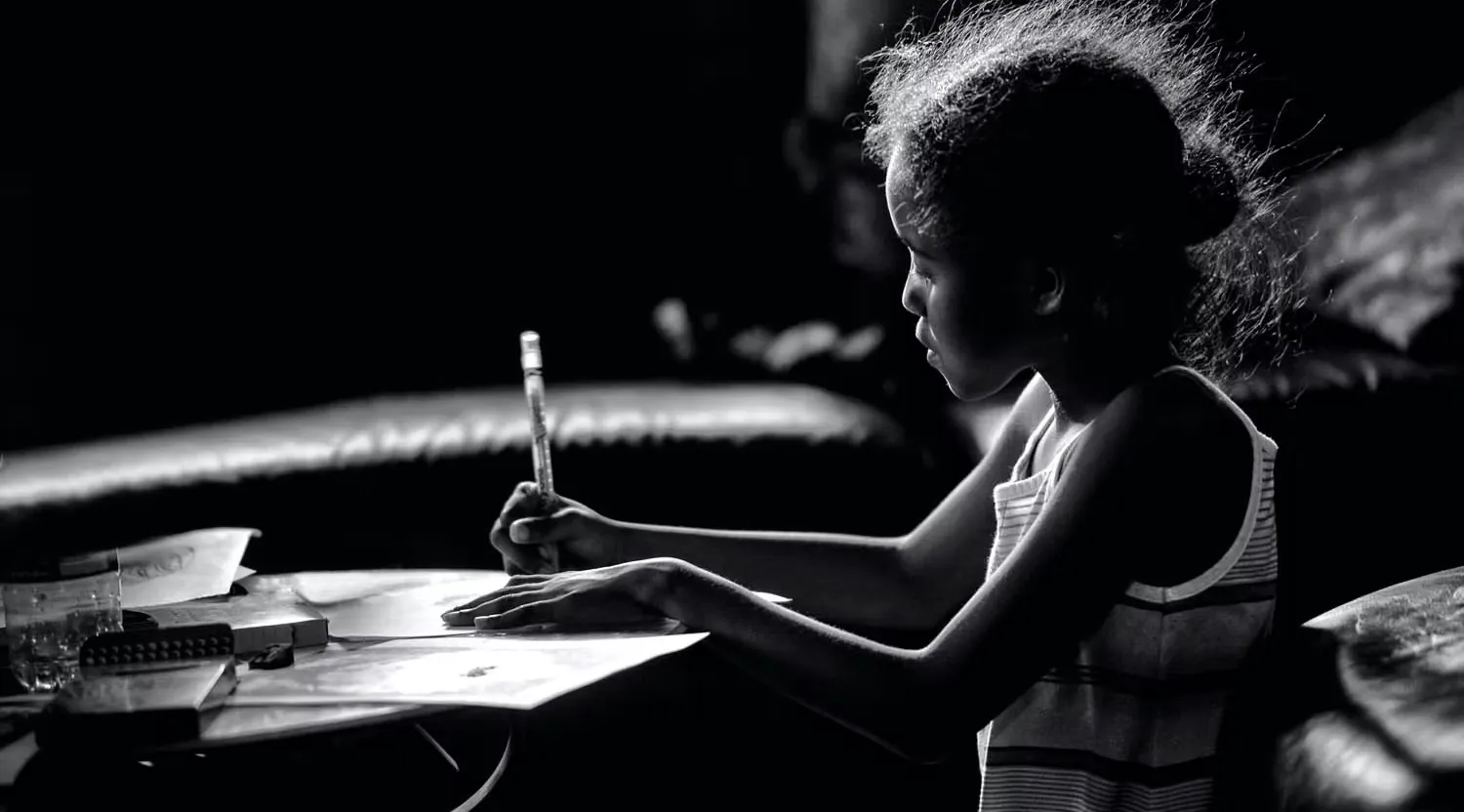 A young girl drawing with a pencil at a table, lit from one side in a darkened room.