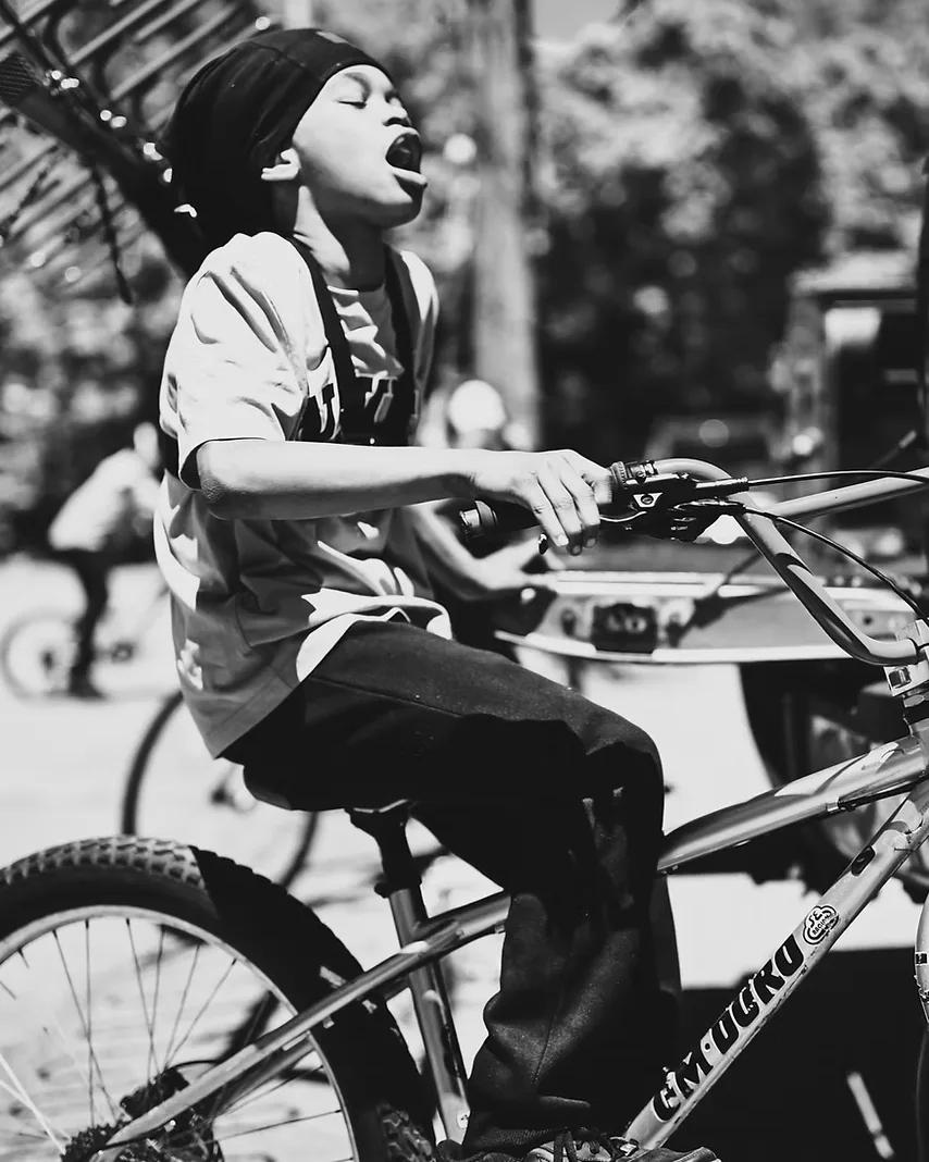 A young rider on a BMX bike at a ride-out, head thrown back mid-shout, other riders behind him.
