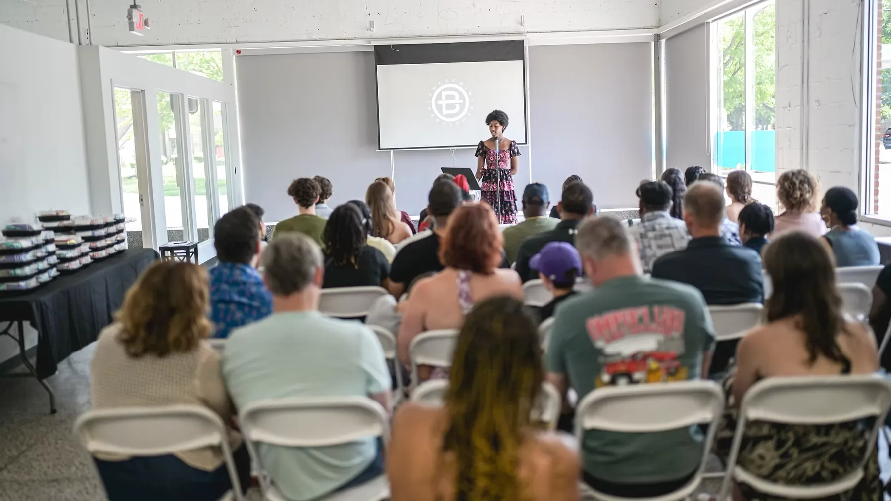 A young woman reads at a microphone to a full room of seated guests, the Balance Point Studios seal projected on the screen behind her.