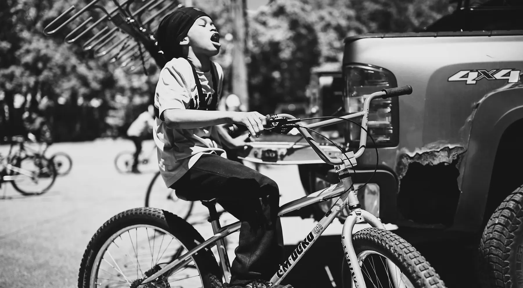 A young rider on a BMX bike at a ride-out, head thrown back mid-shout, other riders behind him.